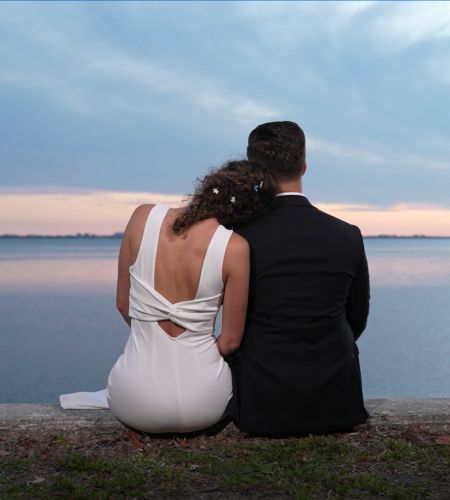 A couple with their backs to the camera sitting on a cliff facing water with the bride's head on the groom's shoulder.