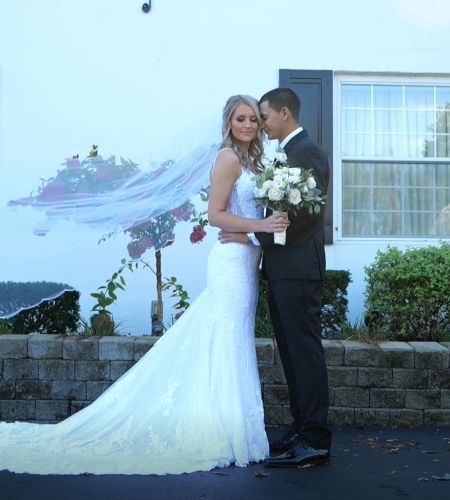 A bride and groom holding each other and standing in front of a mansion with the bride looking directly to camera.