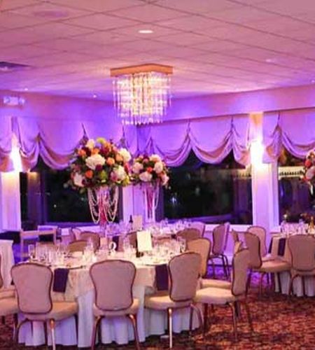 An elegant indoor reception room with lavender lighting, tall floral centerpieces, and a crystal chandelier.